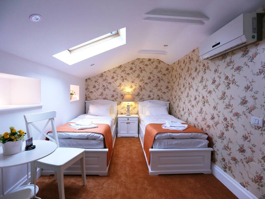 Una habitación pequeña y luminosa con dos camas, una mesa y una silla. Hay un tragaluz sobre las camas y un equipo de aire acondicionado en la pared.