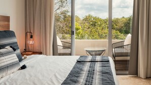 A bedroom with a bed, a nightstand with a lamp, a balcony with a chair, and a view of trees and sky. - NHOA 312B Lux 1BR in Aldea Zama (Tulum)