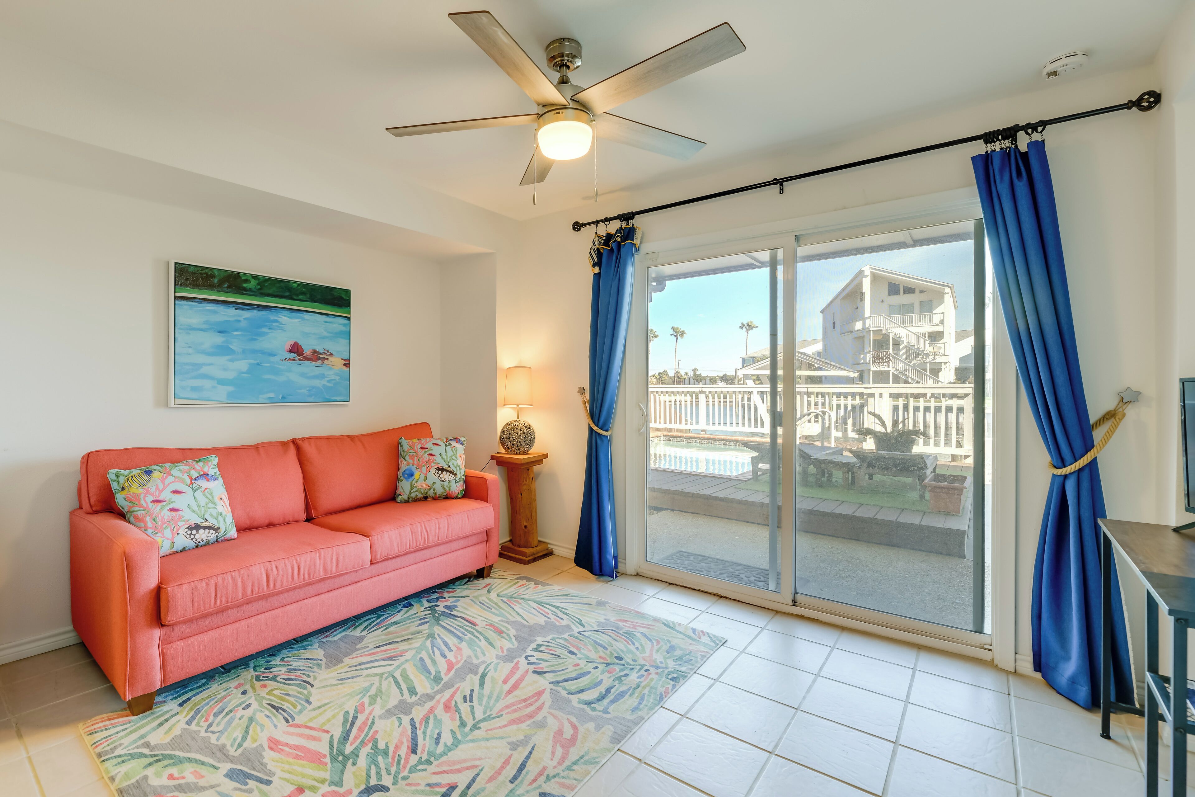 A living room with a pink sofa, a ceiling fan, a large window with blue curtains, and a view of a building outside.