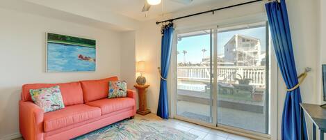 A living room with a pink sofa, a ceiling fan, a large window with blue curtains, and a view of a building outside.