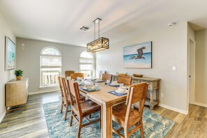Uma sala de jantar com uma mesa de madeira posta para seis pessoas, rodeada de cadeiras, com uma grande janela e um quadro na parede.