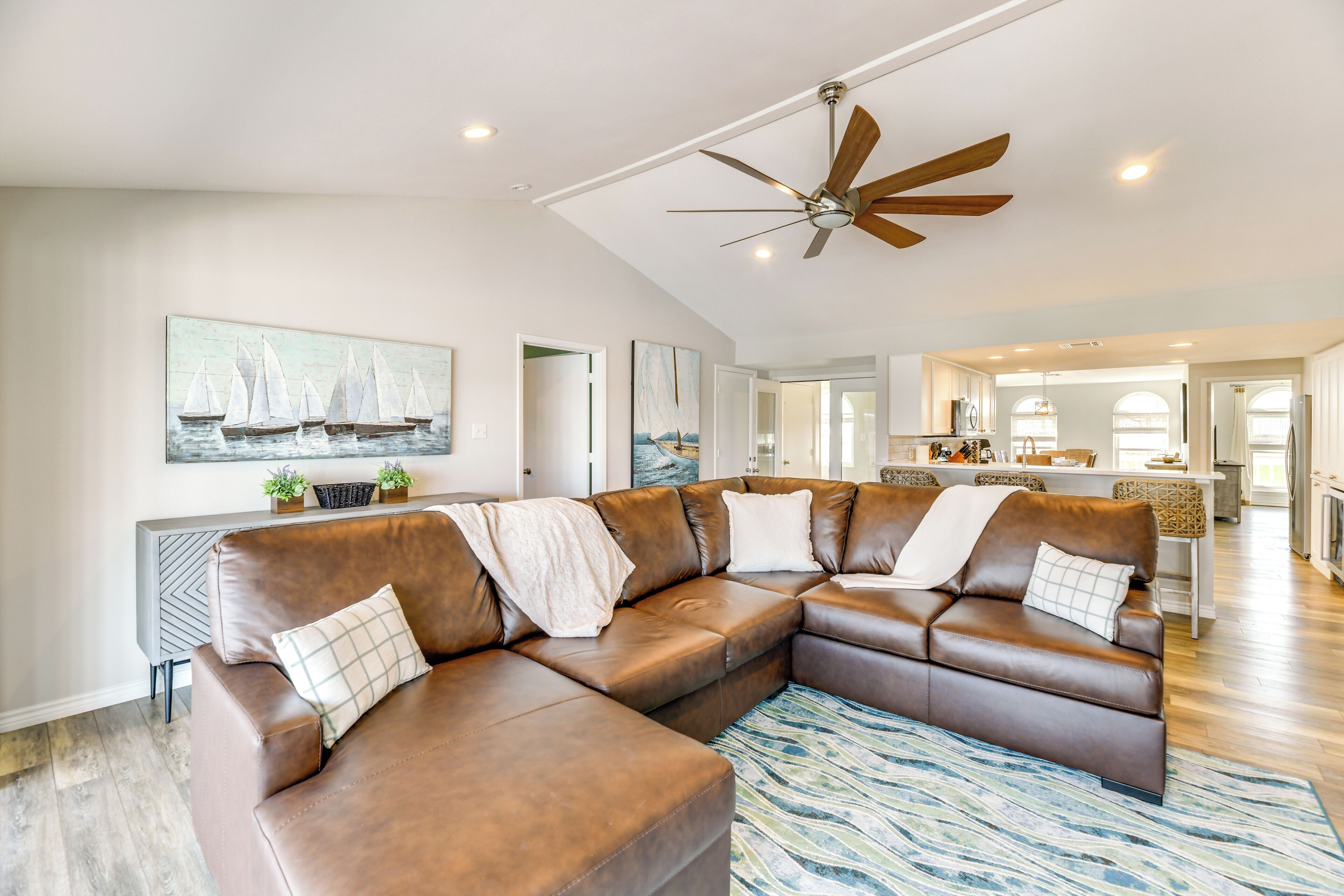 A spacious living room with a large brown leather sectional sofa, a ceiling fan, and a view into the kitchen area.