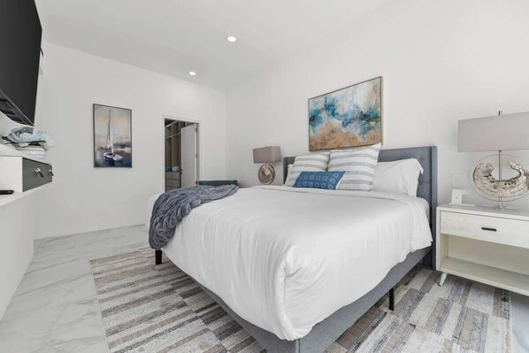 Une chambre à coucher moderne, avec un lit, des tables de chevet, une commode, une œuvre d’art accrochée au mur et une porte.