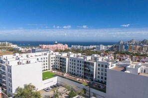 Ein hoher Wohnkomplex mit mehreren Wohnhäusern, einem zentralen Innenhof mit Rasenfläche und Blick auf das Meer sowie entfernte Felsformationen.