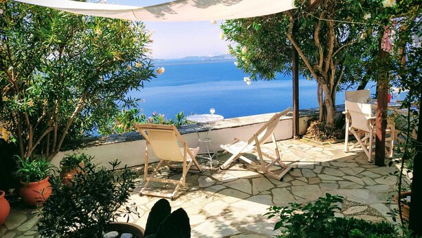 A patio with stone flooring, two wooden chairs, a table, and a view of the sea.