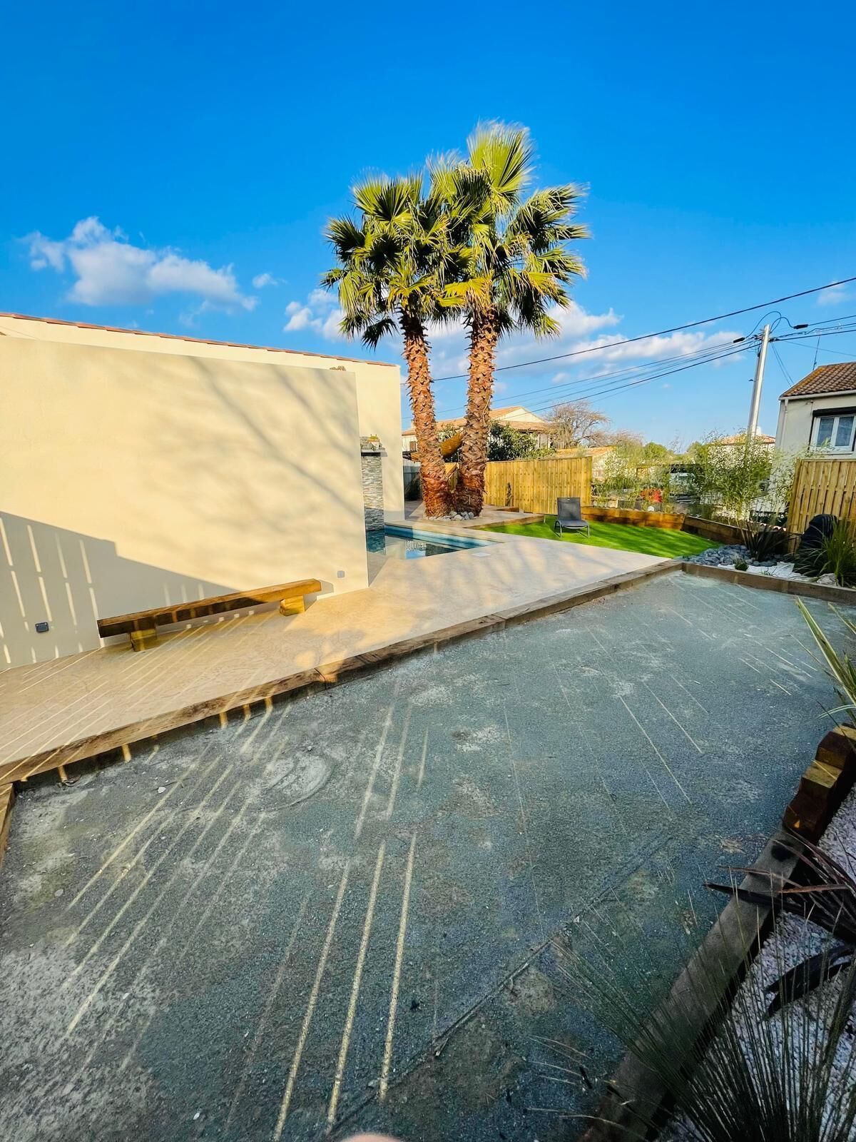 A residential backyard with a swimming pool, two palm trees, a wooden deck, and a fenced area.
