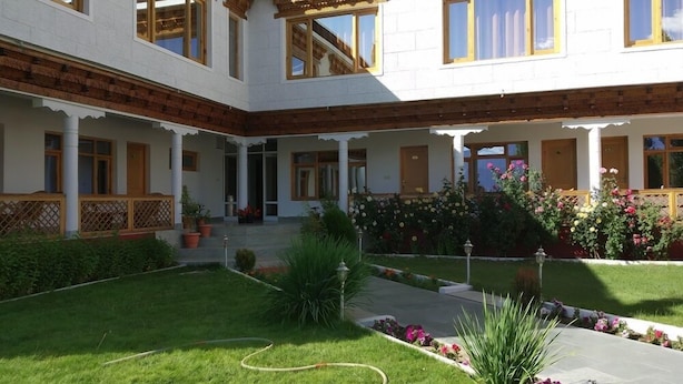 Edificio de dos pisos con un patio interior. La fachada es blanca con detalles de madera y cuenta con múltiples ventanas. En el patio hay un jardín con diversas plantas y flores.