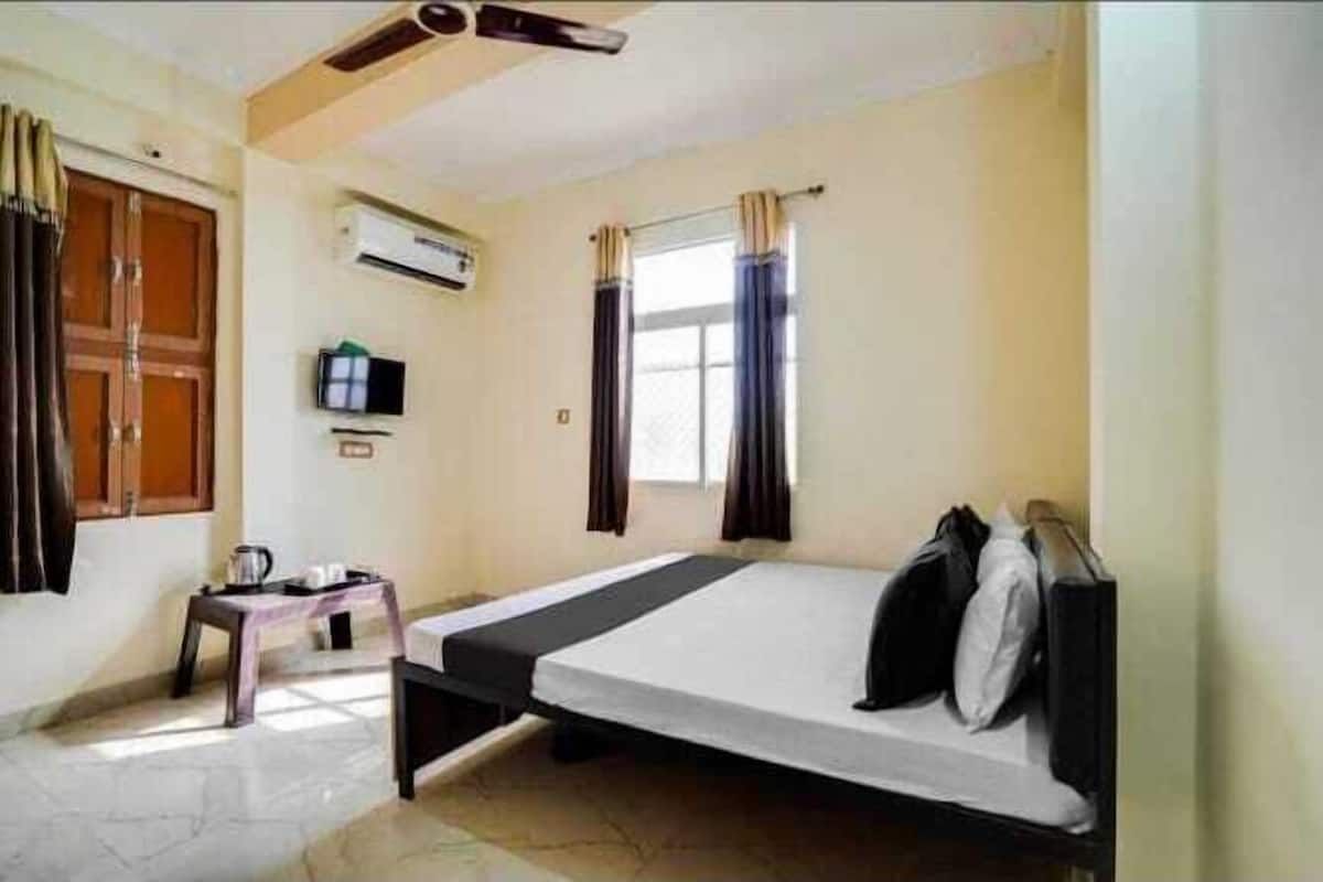 Habitación con cama individual, ventilador de techo, aire acondicionado, una mesita con hervidor y ventana con cortinas.