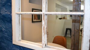 Una ventana con pintura descascarada, un cuadro en la pared y una silla al fondo.