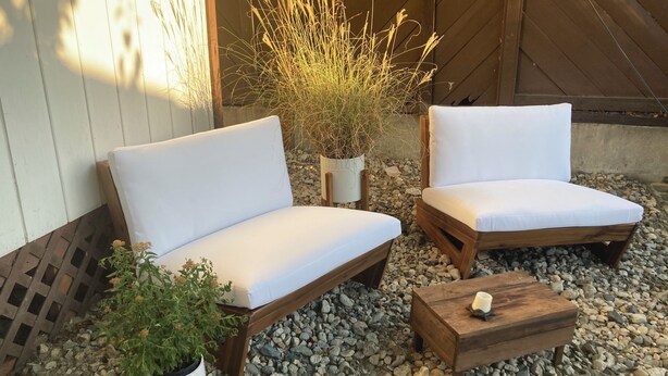 Dos sillas blancas con cojines y estructura de madera, acompañadas de una pequeña mesa de madera con una vela, sobre una superficie de grava con plantas y una valla de madera al fondo.