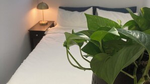 Una cama bien hecha con sábanas blancas, una lámpara de noche y una planta en maceta sobre una mesita de noche.