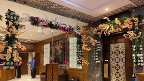A hotel lobby with a reception desk, decorative floral arrangements, and a 'Restaurant' sign.