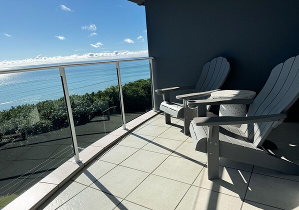 A balcony with a view of the ocean, featuring two Adirondack chairs and a small table.