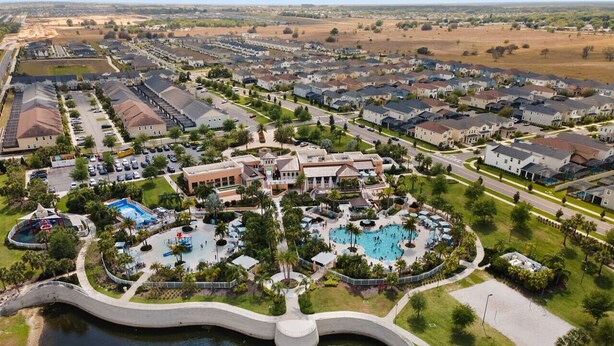 An aerial view of a residential area with a central swimming pool complex surrounded by houses, a bridge over water, and a park area.