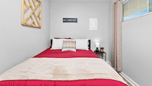 Une chambre à coucher avec un lit, une table de chevet, un panneau fixé au mur et une fenêtre avec des rideaux.