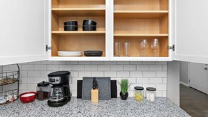 Une cuisine avec des meubles blancs, un plan de travail en granit, une machine à café, un bloc à couteaux et une plante en pot.