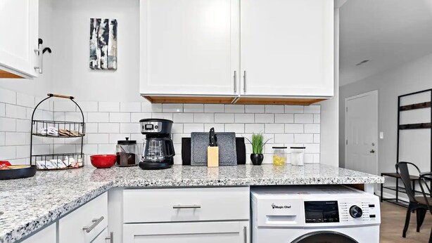 Une cuisine moderne avec des meubles blancs, un plan de travail en granit, une machine à café, un bloc à couteaux et un lave-linge.