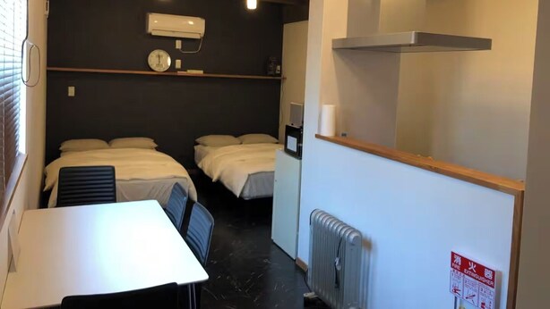A small, compact room with two single beds, a table, and a chair. There is a radiator, a wall-mounted air conditioner, and a ceiling light fixture. - GoodHome Toyokawa (Hakodate)