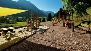 A playground with a slide and climbing structures, surrounded by a gravel area and a yellow shade sail. - Apartment in Kleinarl near Ski Slopes (Kleinarl)