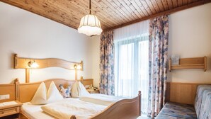 Un lit en bois avec une tête de lit, une table de chevet et une lampe. Il y a une fenêtre avec des rideaux à motifs floraux et un banc avec un coussin à motifs.