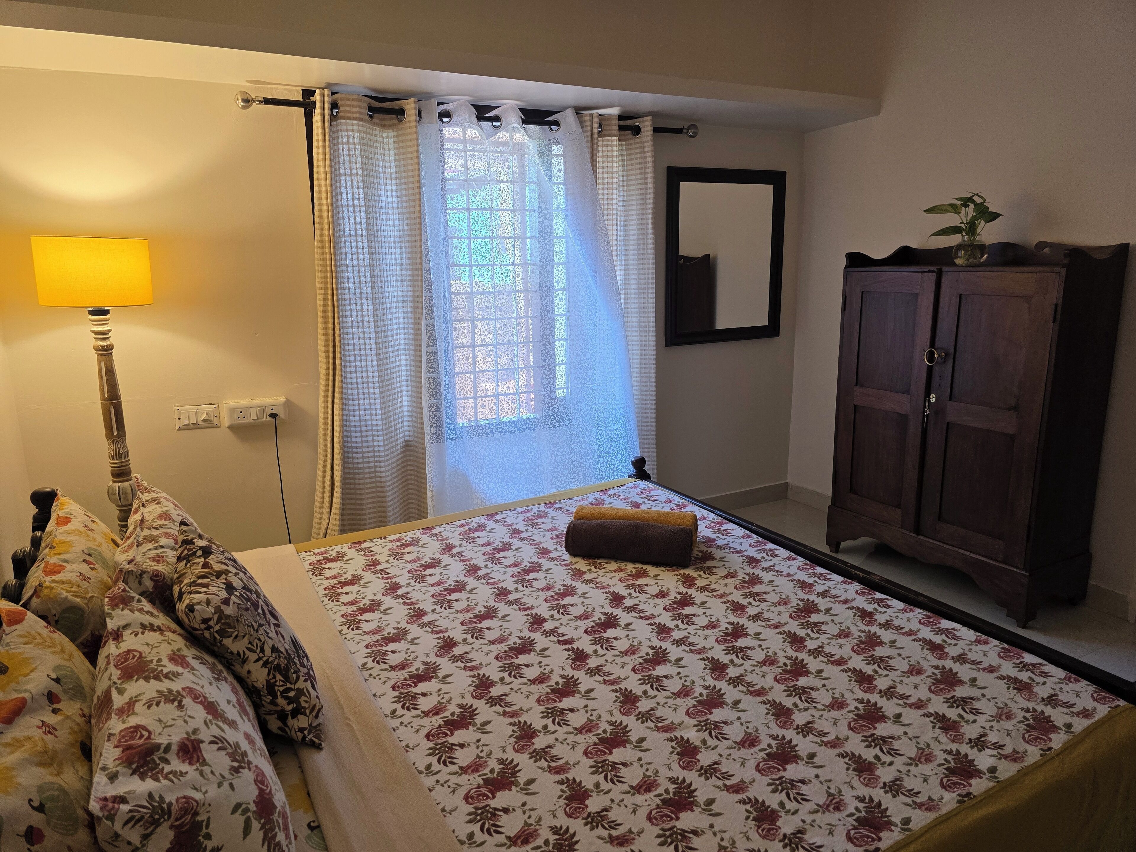 A bedroom with a bed, a nightstand, a lamp, a window with curtains, a mirror, and a wardrobe.