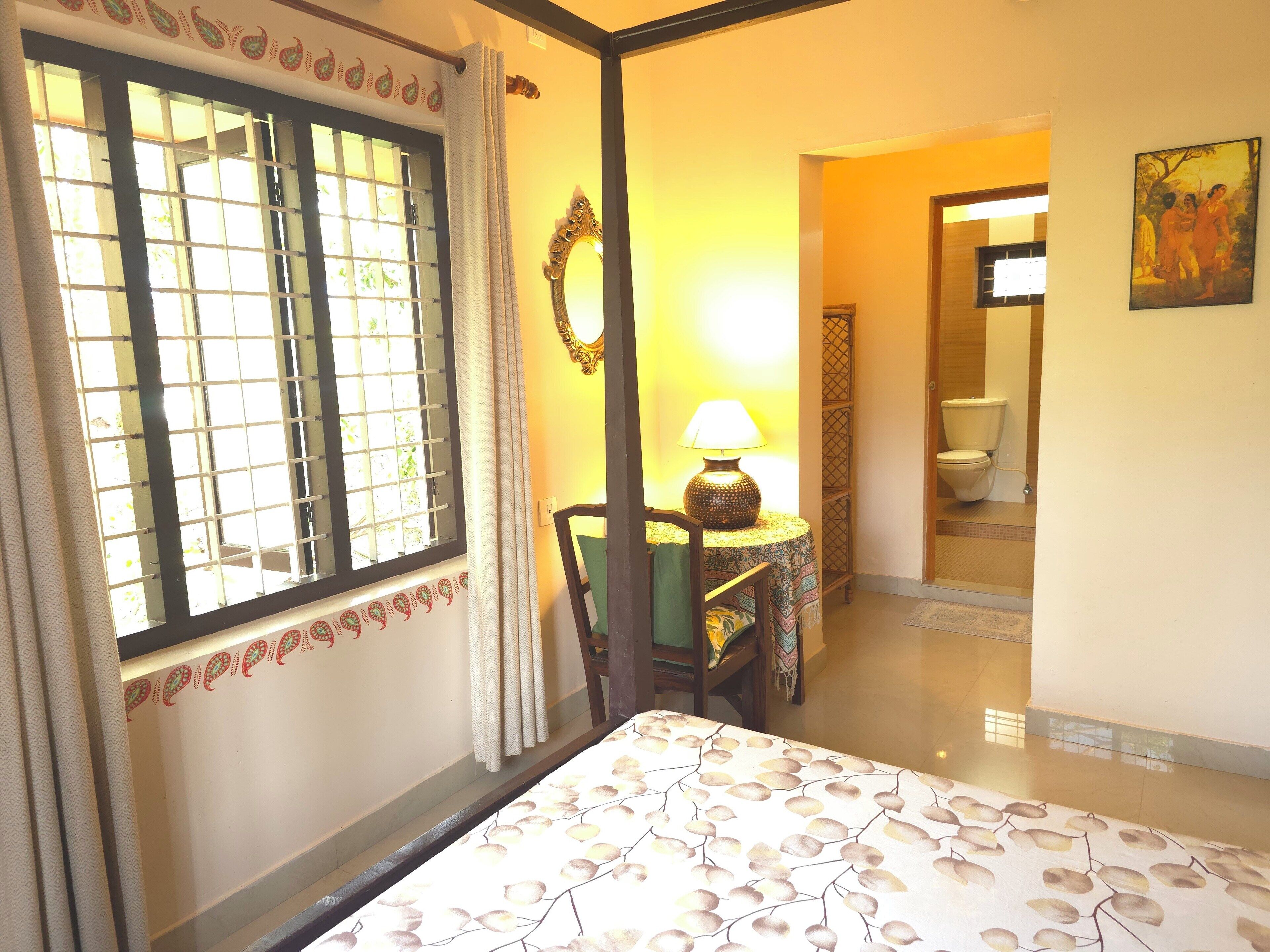 A hotel room with a bed, a table with a lamp, a chair, a window with a curtain, and a painting on the wall.