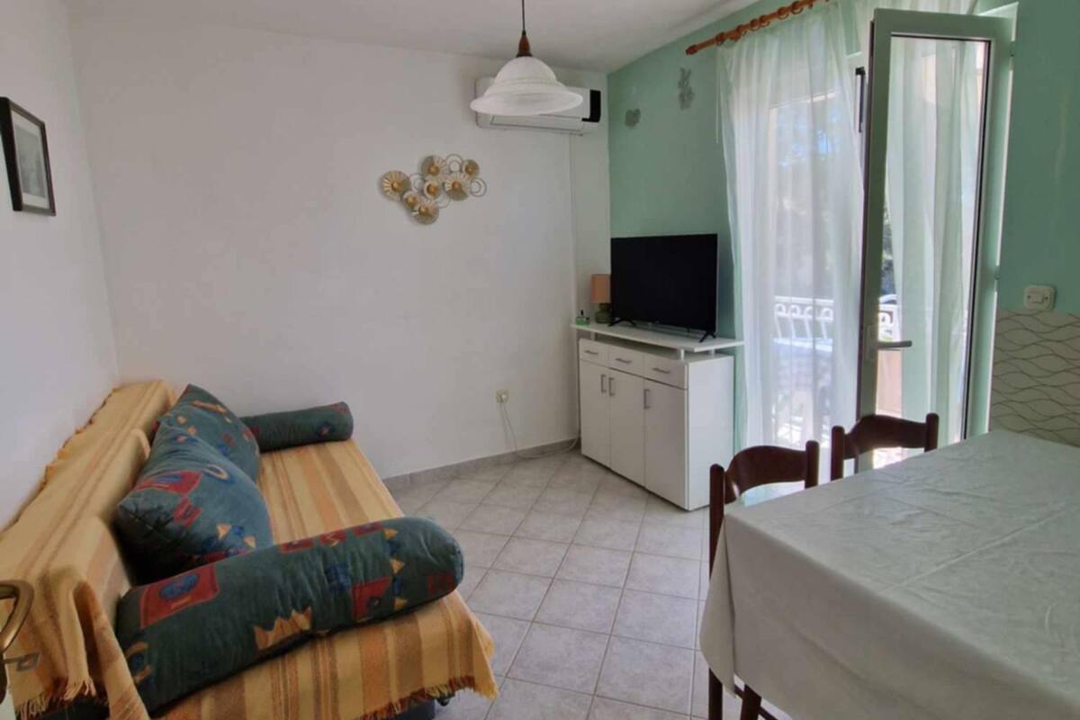 A small, simple room with a sofa, a TV on a cabinet, a dining table, and a window with curtains.