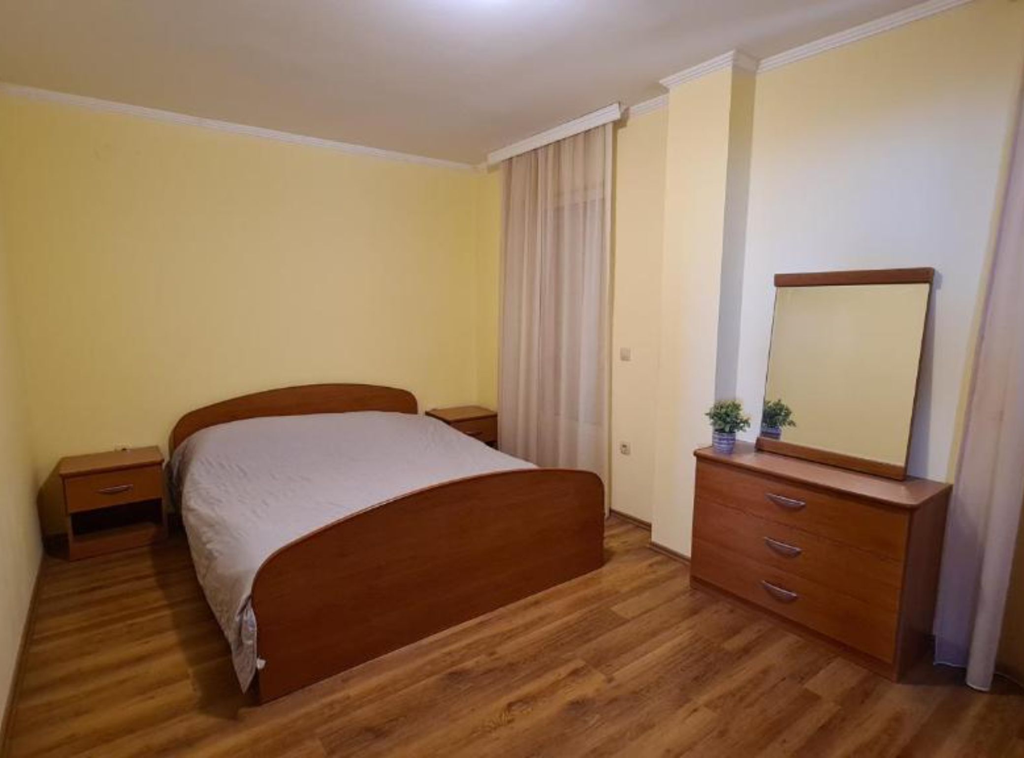 Une chambre à coucher avec un lit en bois, une table de chevet, une commode avec un miroir et une fenêtre avec des rideaux.