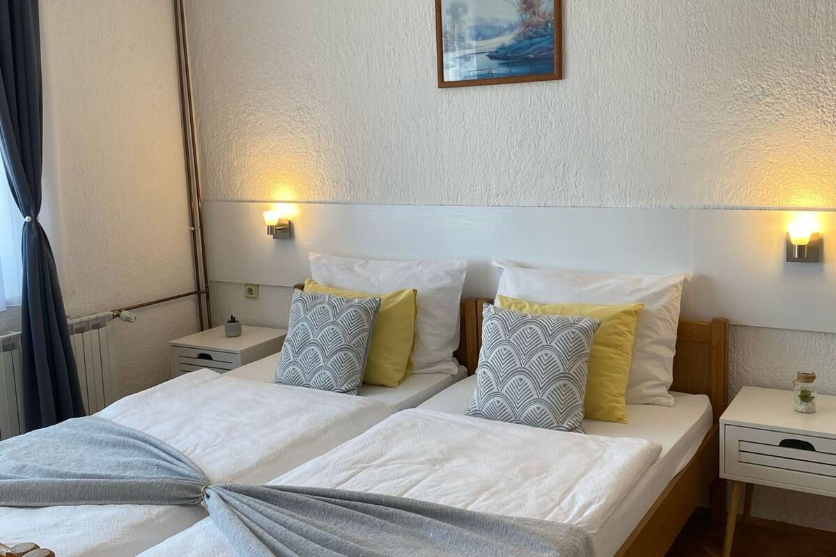 A double bed with two pillows, a bedside table, a nightstand, a wall-mounted picture, and a window with curtains.