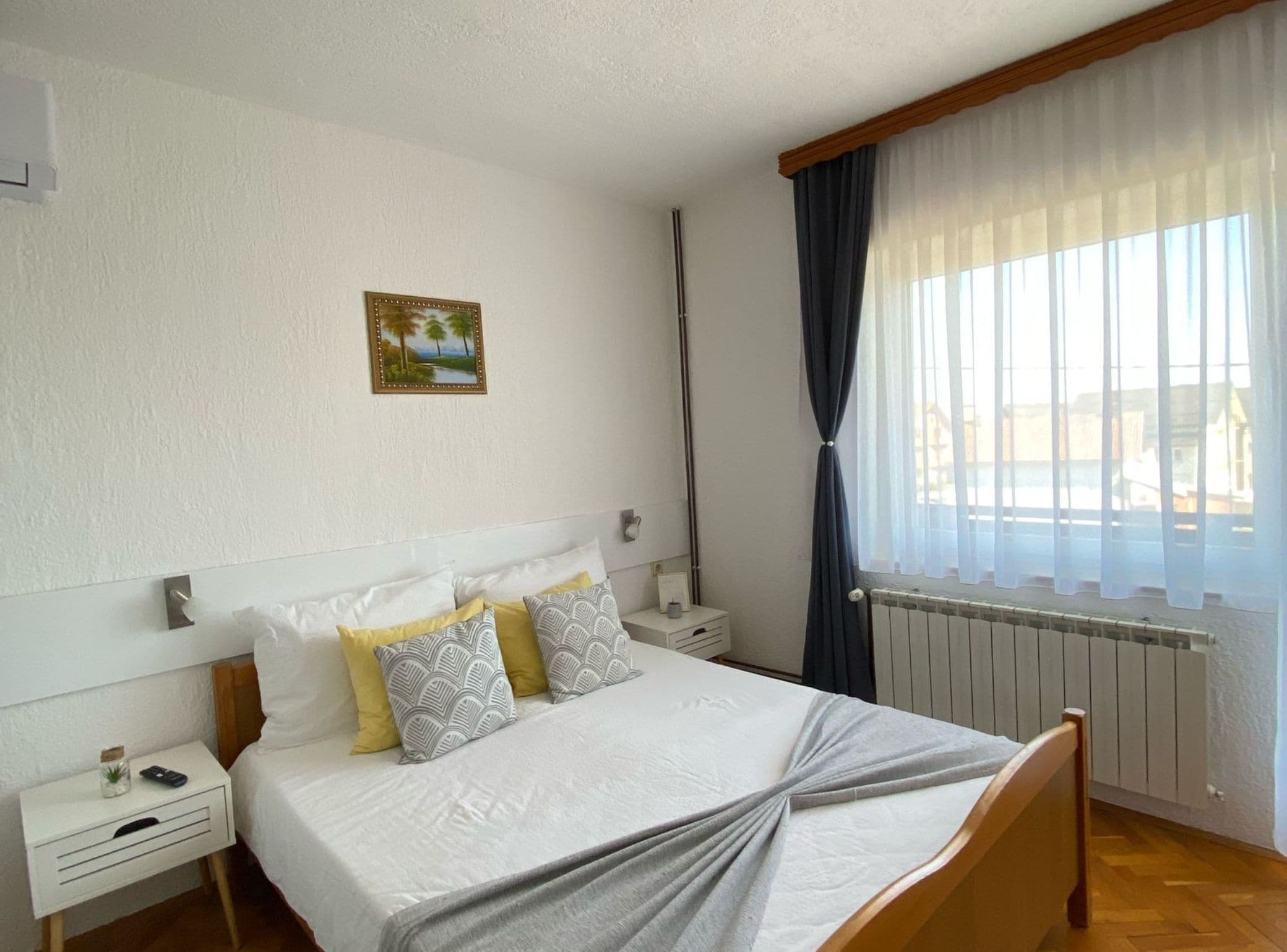 A neatly arranged bedroom with a bed, bedside tables, a nightstand, a picture on the wall, and a window with curtains.