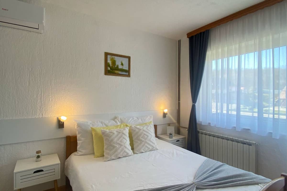 A neatly arranged hotel room with a bed, bedside tables, a nightstand, a picture on the wall, and a window with curtains.