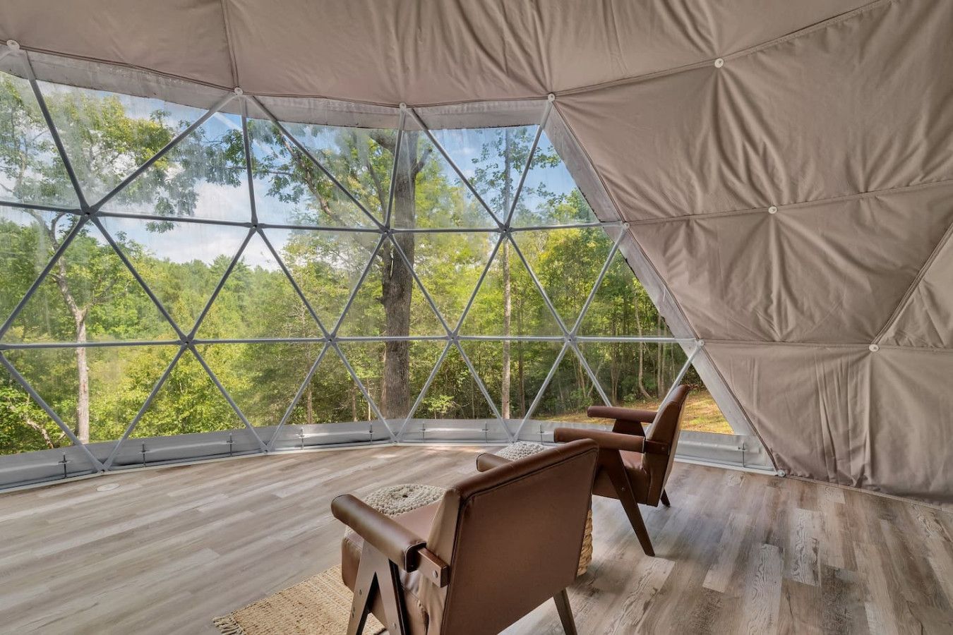 A geodesic dome room with a wooden floor and two leather chairs, offering a view of a forest.