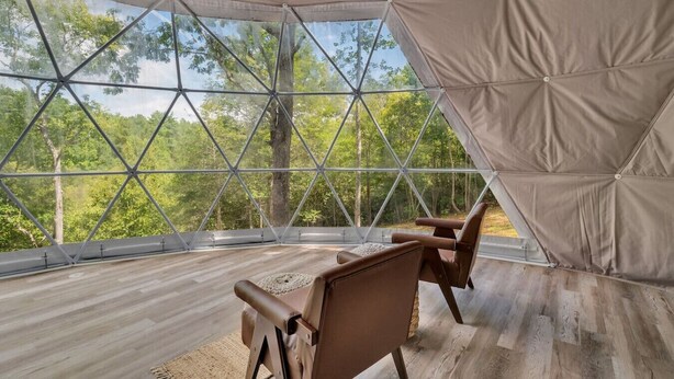A geodesic dome room with a wooden floor and two leather chairs, offering a view of a forest. - Nottely River Retreat | Fire Pit (Murphy)