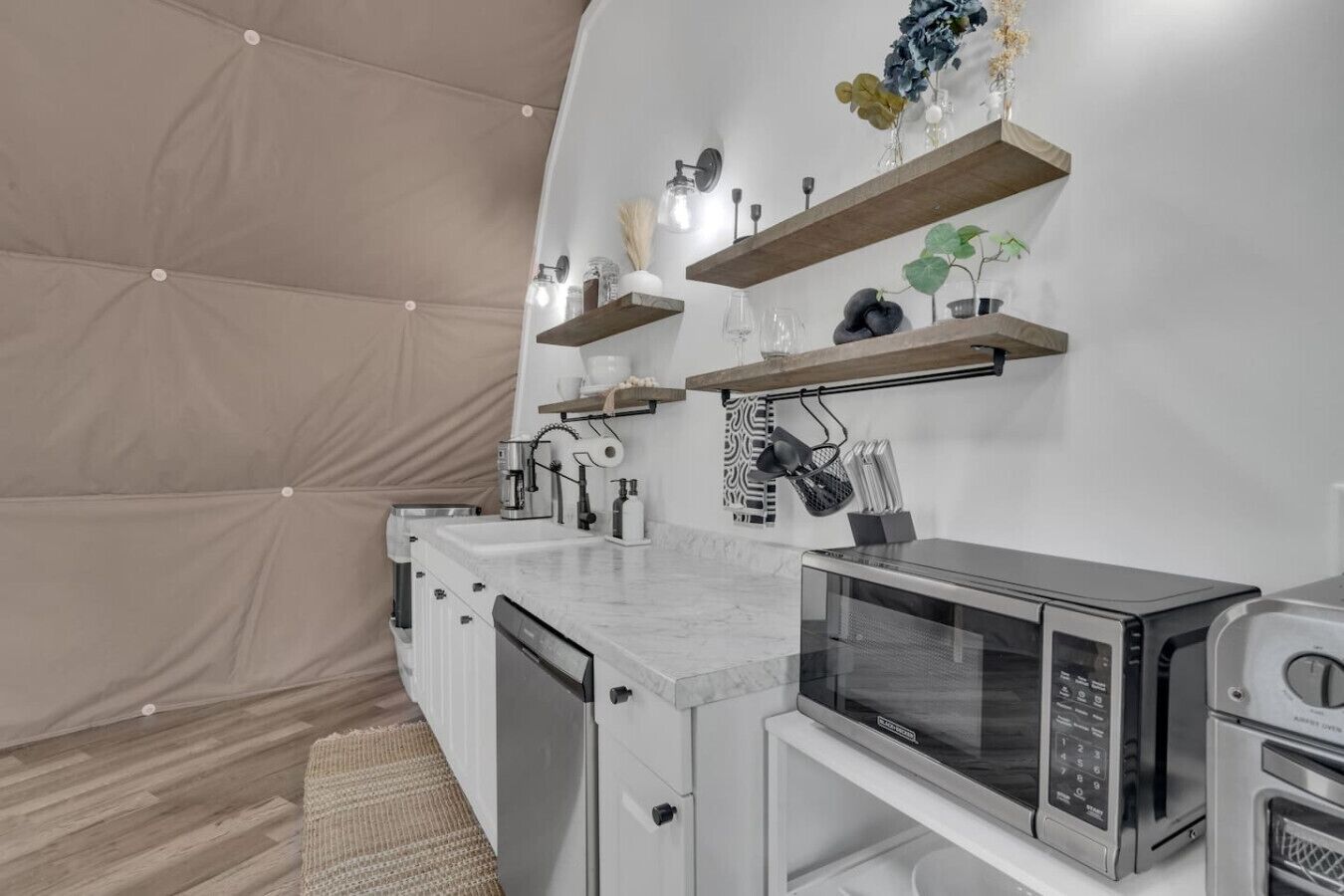 A compact kitchen with a microwave, toaster, and coffee maker. There are wooden shelves above the counter holding various kitchen items.