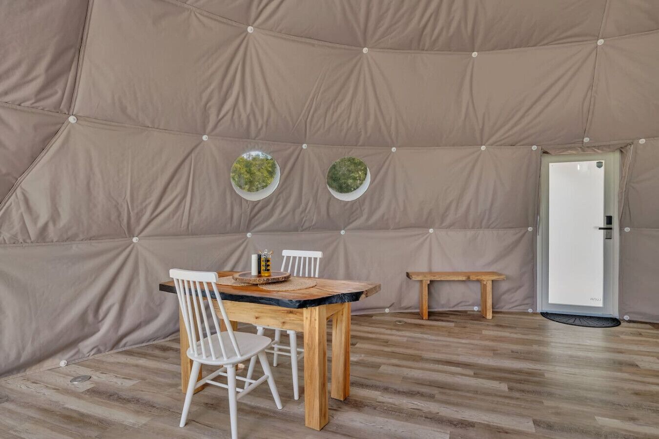 Una habitación tipo tienda de campaña con una mesa de madera y sillas, dos ventanas circulares pequeñas y una puerta blanca.