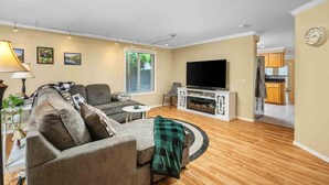 A living room with a sofa, TV, and a small table. There is a window with blinds, a lamp, and a plant. - Fantastic Single Level Ranch Layout, Fenced Backyard w/ Large Deck, New Decor and Furnishing (Hillsboro)