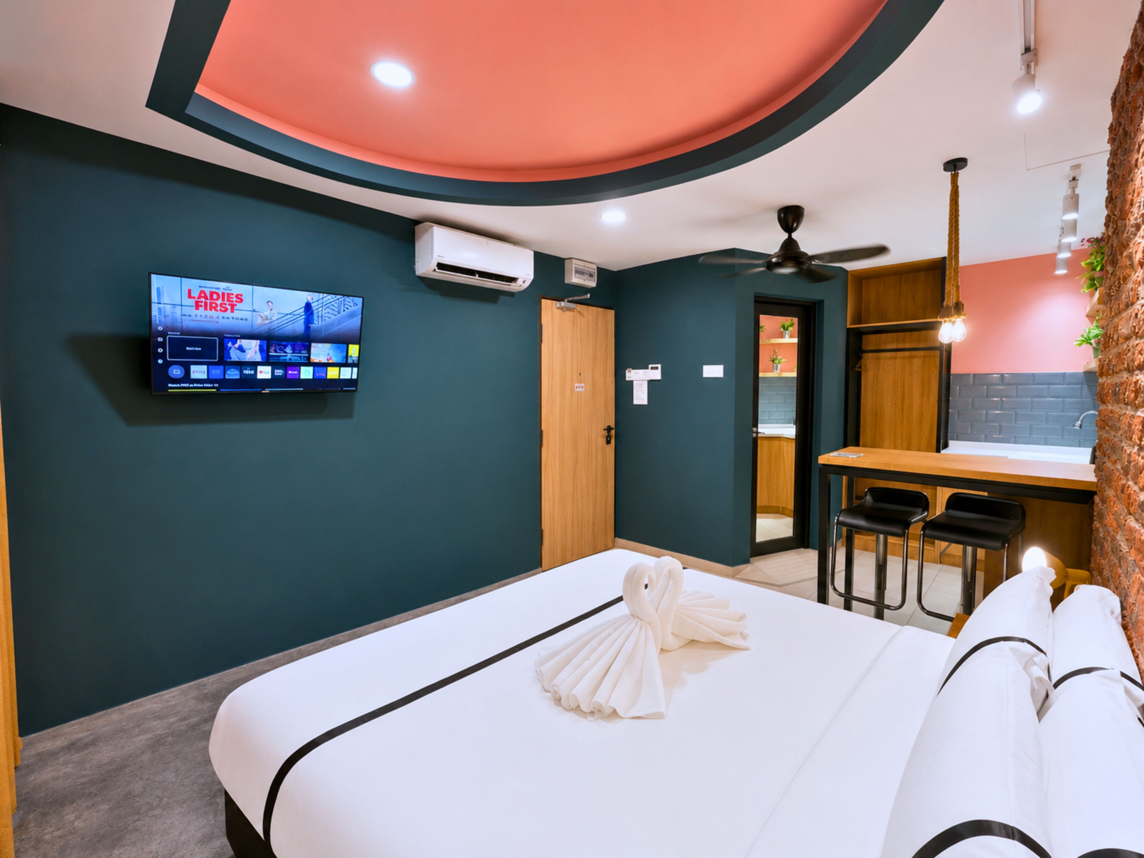 A hotel room with a bed, a TV mounted on the wall, a desk with chairs, and a ceiling fan.