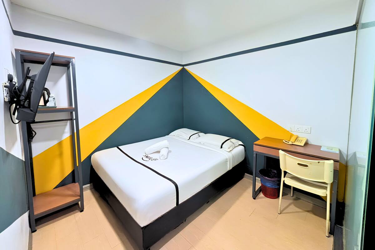 Un lit simple avec un matelas et des oreillers blancs, un bureau avec un téléphone, une chaise et une petite poubelle.