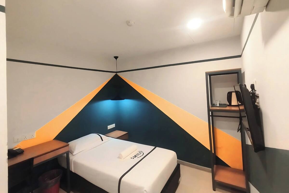 Une chambre d’hôtel de taille réduite, comprenant un lit, un bureau, une chaise, une petite table, une poubelle et un miroir.