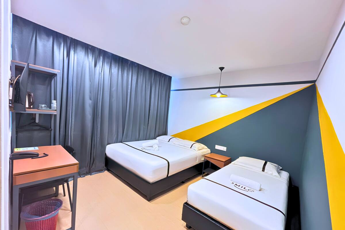 Une chambre d’hôtel compacte, équipée de deux lits, d’un bureau, d’une étagère avec des articles de toilette et d’un luminaire suspendu.