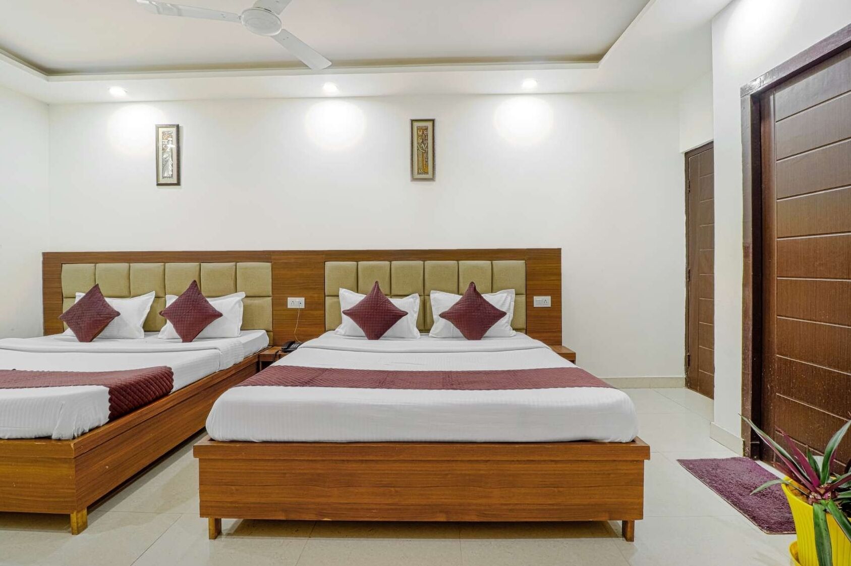 A hotel room with two beds, each with a wooden headboard, white bedding, and maroon pillows. There are ceiling lights and a ceiling fan.