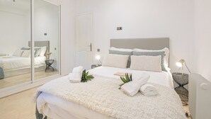 Una cama bien tendida con una colcha blanca y almohadas, una mesita de noche con una lámpara y un espejo grande en la pared.
