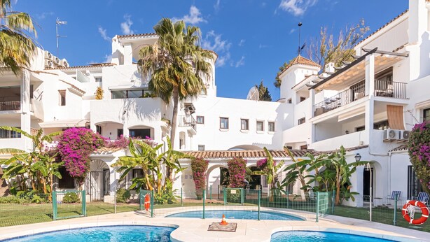 A swimming pool surrounded by white buildings with balconies, palm trees, and purple flowers. - Puerto Banus Near La Sala By The Sea (Marbella)