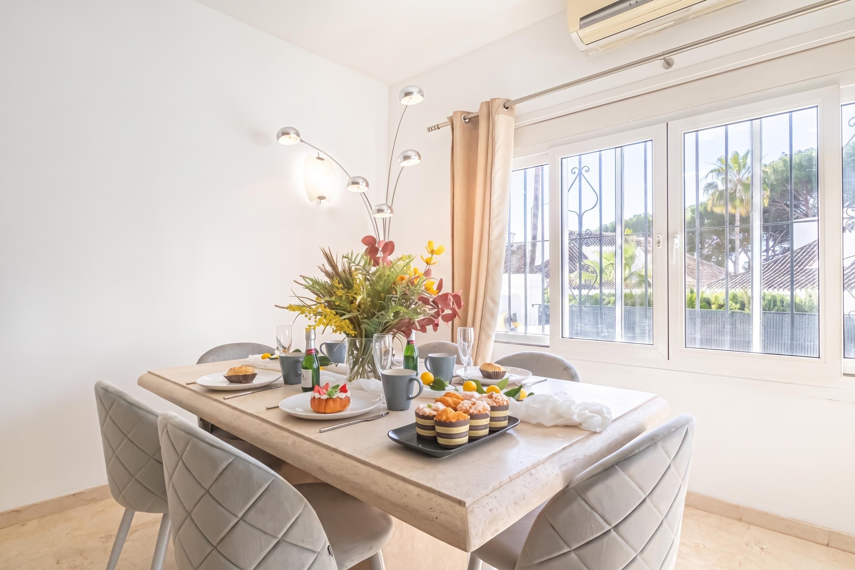 Comedor con una mesa dispuesta para dos personas, un centro de mesa floral, una variedad de pasteles y vistas al exterior a través de grandes ventanales.