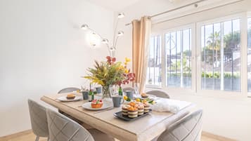 Comedor con una mesa dispuesta para dos personas, un centro de mesa floral, una variedad de pasteles y vistas al exterior a través de grandes ventanales.