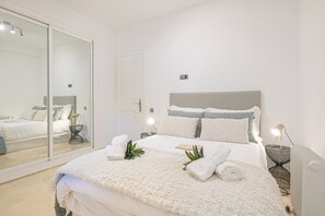 A neatly made bed with a white comforter and pillows, a bedside table with a lamp, and a large mirror on the wall.