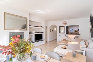 Ein modernes Wohnzimmer mit Fernseher, eine Essecke mit Tisch für zwei Personen und eine Küchenzeile mit Mikrowelle und Kaffeemaschine.