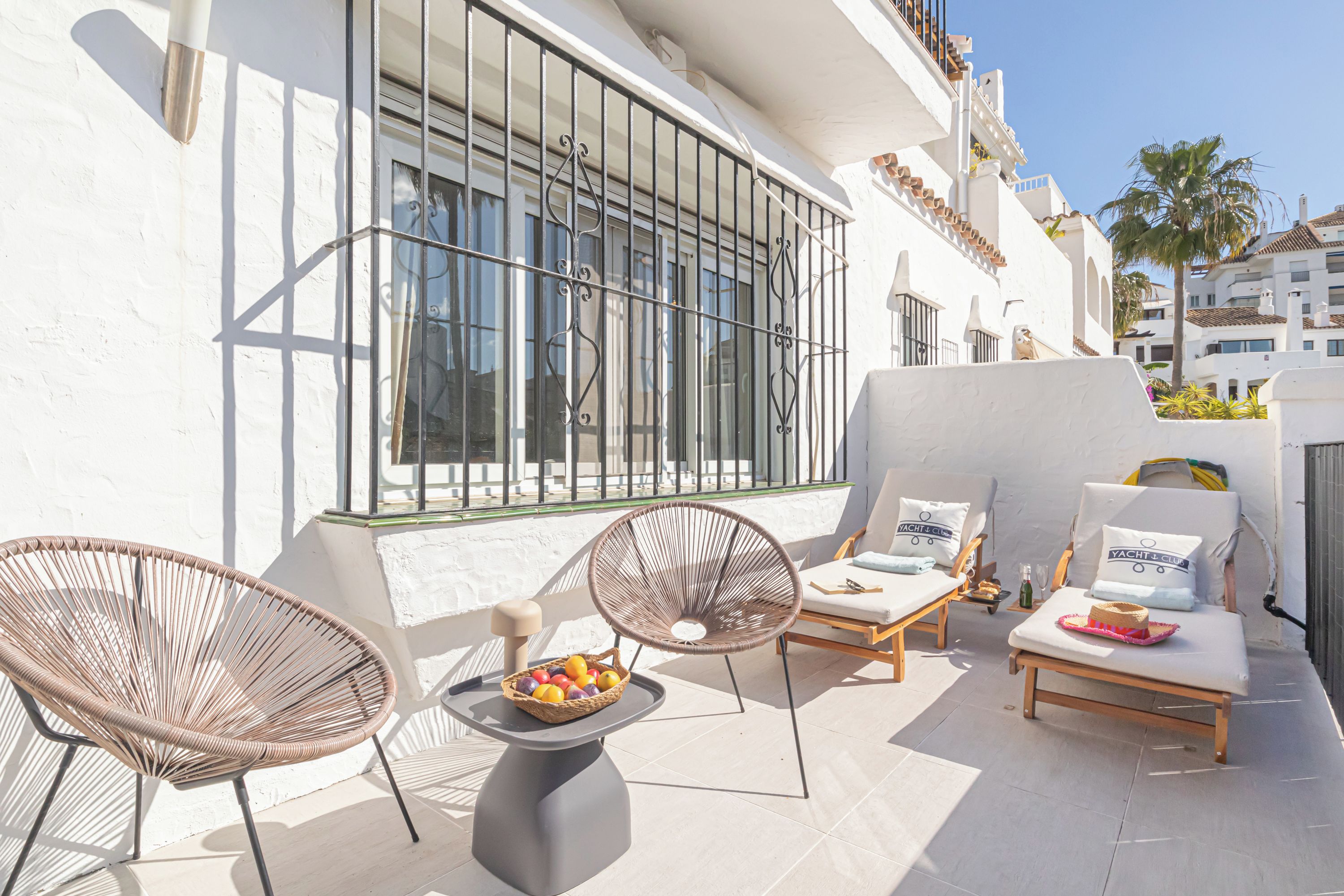 Un espacio al aire libre con luz solar, sillas de mimbre, una mesa con fruta y una silla lounge con un cojín que dice 'YACHT CLUB'.