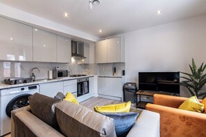 A modern kitchen with built-in appliances, a dining area with a sofa and cushions, and a flat-screen TV. - Central 2BR Apt Basingstoke Near Shops & Transport (Basingstoke)