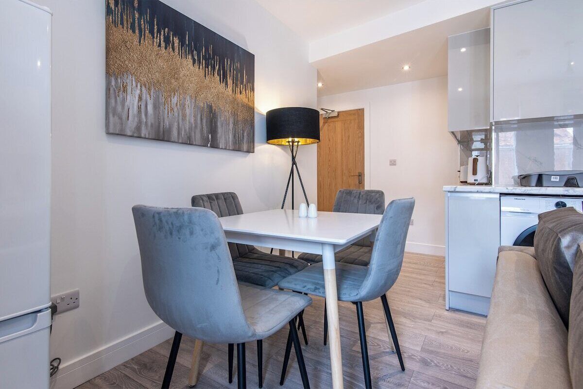 A modern dining area with a white table and grey upholstered chairs, a black floor lamp, and a kitchen in the background.